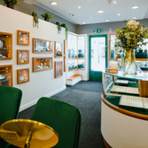 The bright and welcoming interior of the Griffin & Co showroom. The view shows a curved glass counter, green velvet consultation chairs, and jewellery displayed in wood-framed cases on the wall, with the entrance door in the distance.